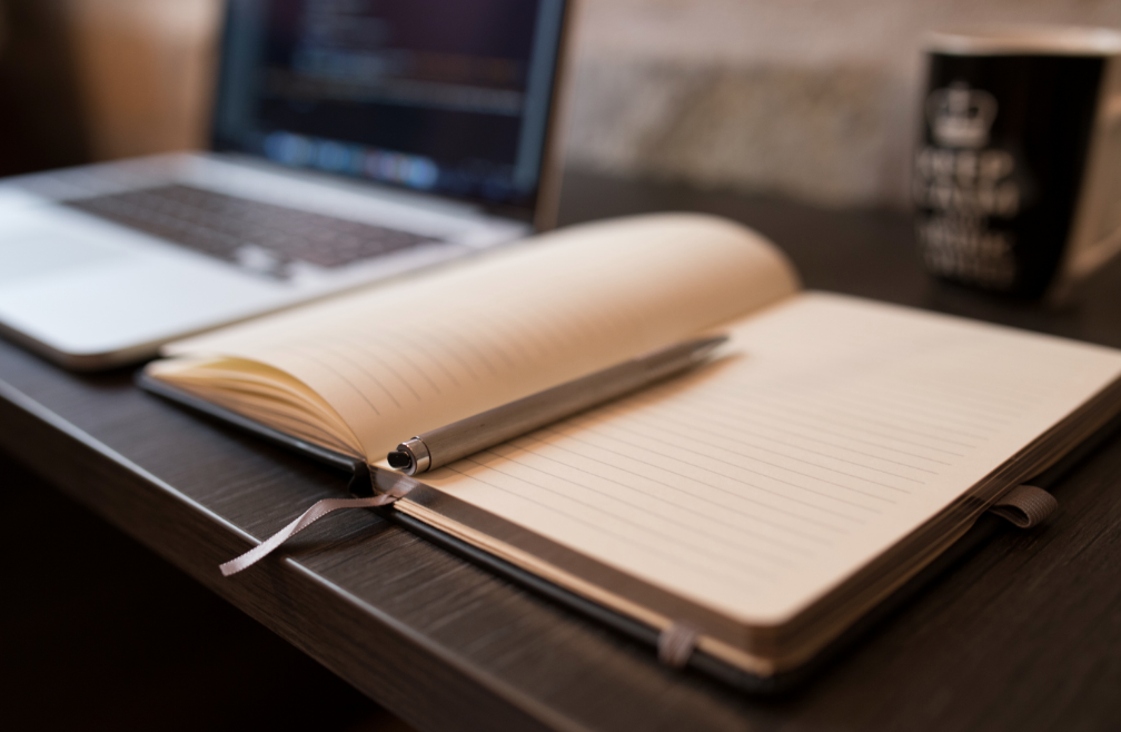 Open empty paper notebook with blurry laptop in the background.